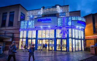 Light up blue ribbon bow with bright white curtain lights and bright white snowflake motifs on the outside wall of Alhambra Shopping Centre.