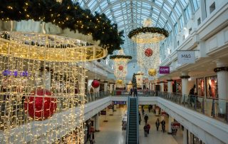 A Christmas display at Royal Priors Shopping Centre including warm white curtain light chandeliers suspended from the ceiling with green light up garlands placed around the top of them and red and gold baubles place in the centre of them.