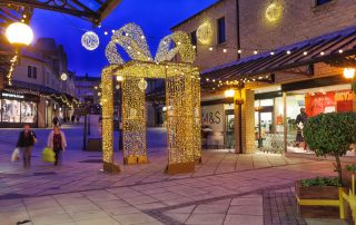 Light up gold present box displayed outdoors with warm white aurora balls and festoon lights suspended above it at Woolshops Shopping Centre.