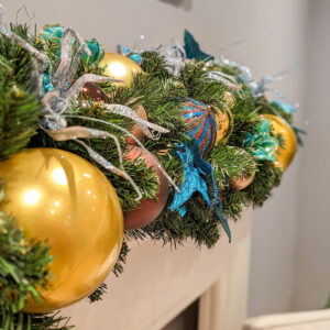 A close up image of an artificial green garland decorated with gold, brown, and blue baubles and floral picks.