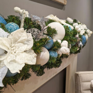 A close up image of an artificial green garland decorated with blue and white baubles and floral picks.