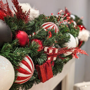 A close up image of an artificial green garland decorated with red, white, and black baubles and floral picks.