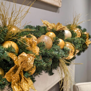 A close up image of an artificial green garland decorated with gold baubles and floral picks.