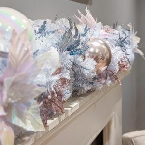 A close up image of an artificial iridescent white garland decorated with iridescent, white, pink, and blue baubles and floral picks.