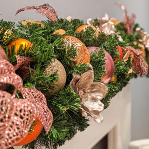 A close up image of an artificial green garland decorated with pink, orange, and gold baubles and floral picks.