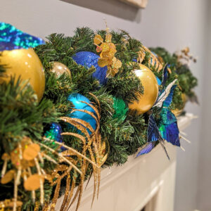 A close up image of an artificial green garland decorated with blue, green, and gold baubles and floral picks.