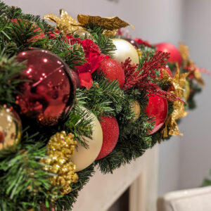 A close up image of an artificial green garland decorated with red and gold baubles and floral picks.